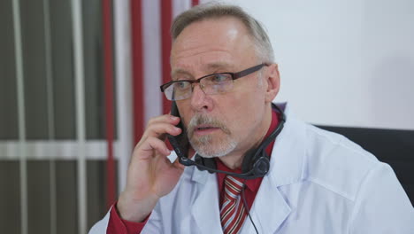 doctor sitting at desk behind his laptop and talking on mobile phone with his patient