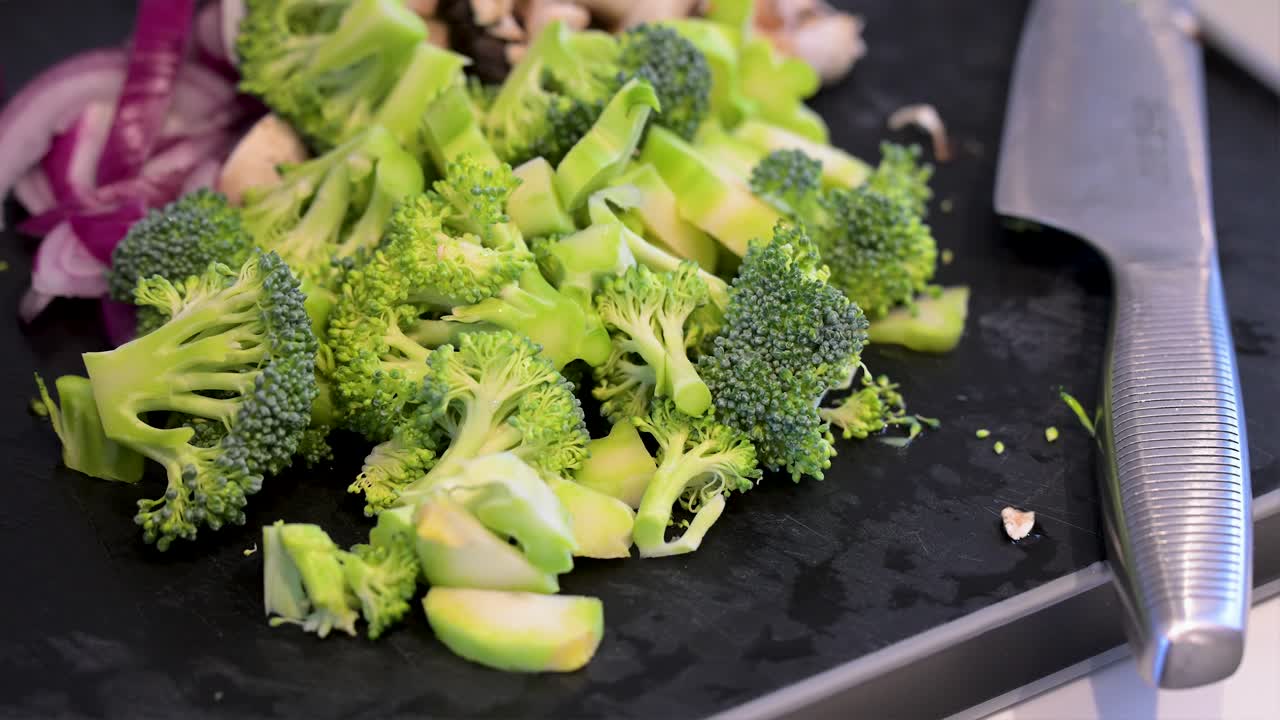 Freshly chopped broccoli and sliced red onions are prepped on a black cutting board next to a chef's knife. A close-up scene of healthy meal preparation and clean eating
