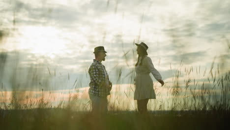 a tender moment between a couple as they embrace warmly in a grass field at sunset. the woman wears a flowing white dress, while the man is in a casual checkered shirt and hat