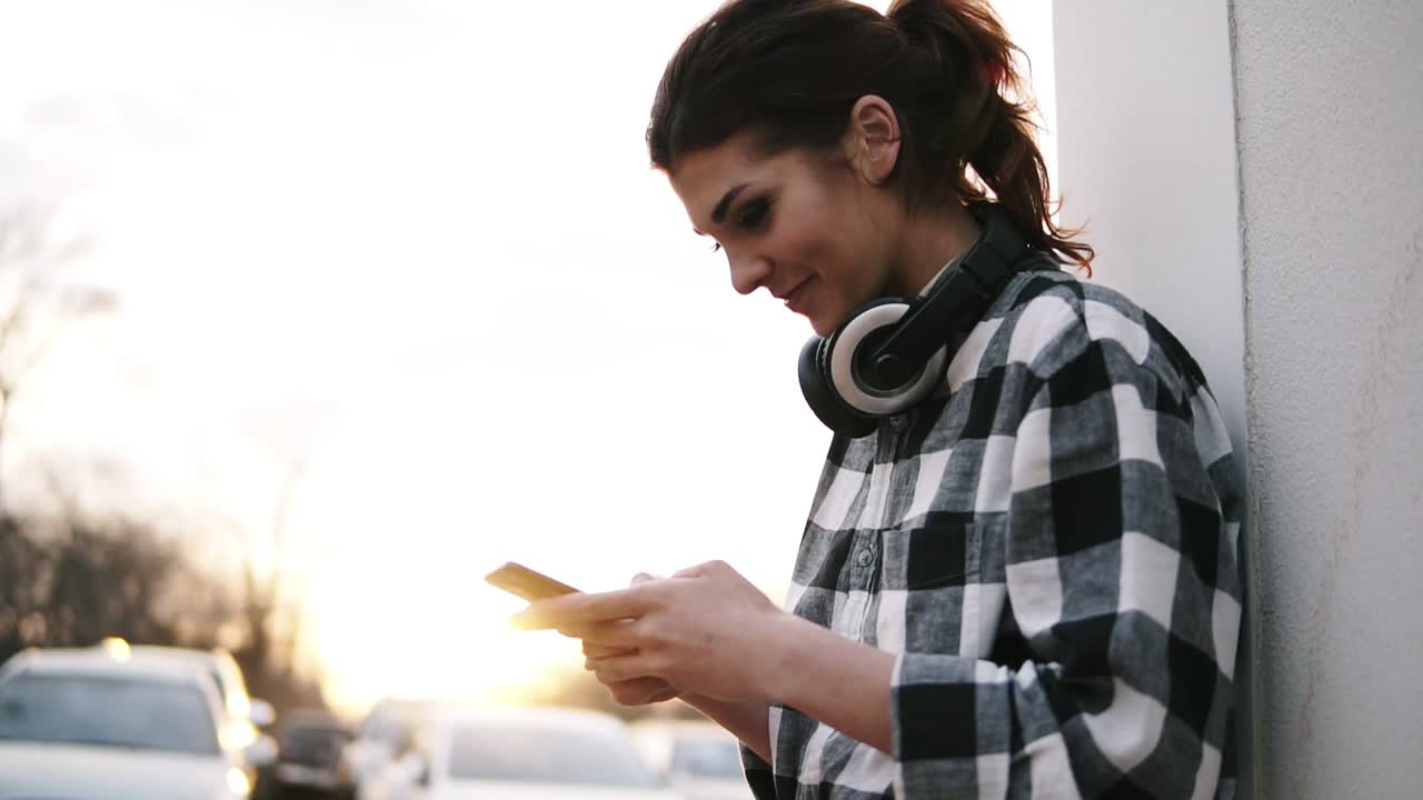 Pretty woman is standing near the white column, holding mobile in her hands and typing. Headphones. The sun is shining