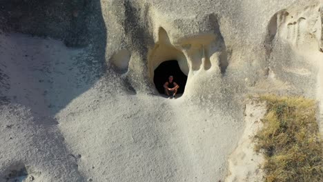 Male-traveler-sitting-in-cave-opening-in-Cappadocia-landscape,-pull-back-aerial