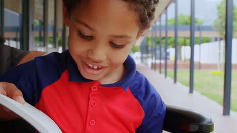 Front-view-of-disabled-African-American-schoolboy-reading-a-book-on-wheelchair-in-school-corridor-4k