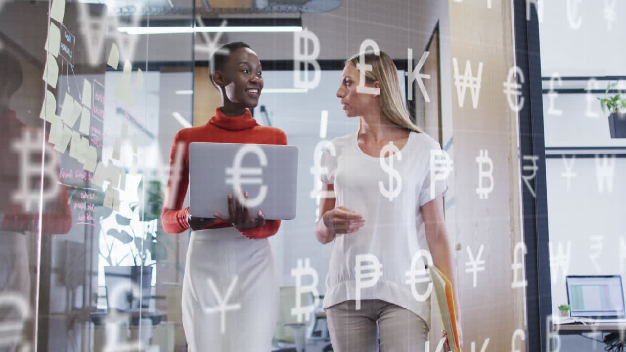 animación de símbolos de moneda contra dos mujeres diversas discutiendo sobre una computadora portátil en la oficina