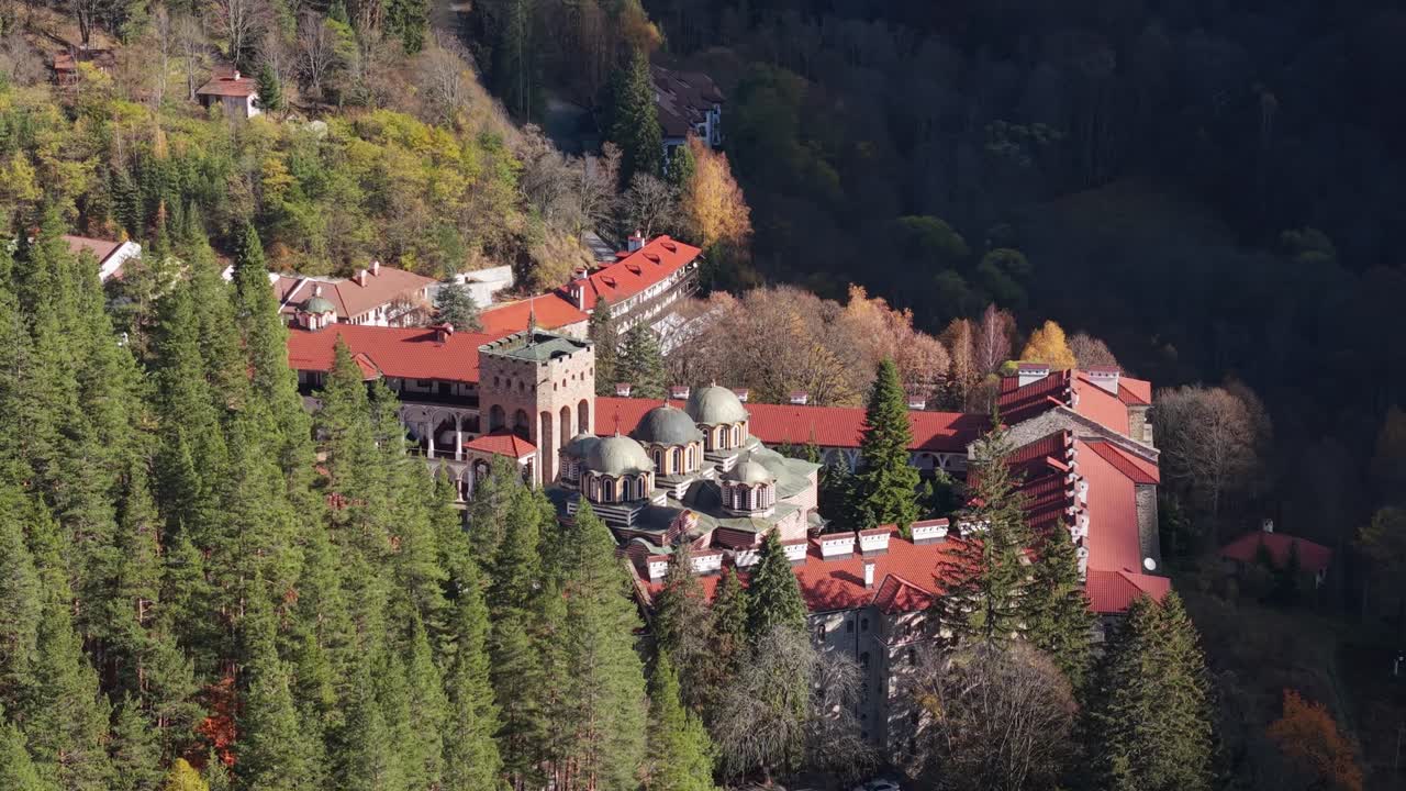 A drone using a telephoto lens gradually reveals Rila Monastery hidden behind trees, capturing the monastery and surrounding forested landscape