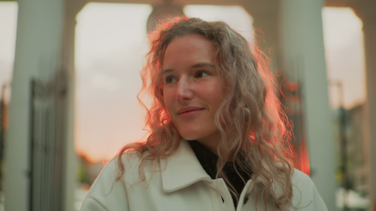 portrait view of fashion designer posing outdoors with warm gentle smile wearing light coat standing against backdrop of white pillars and sunset sky