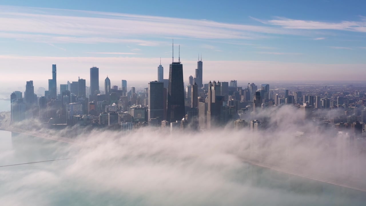Sweeping aerial footage reveals Chicago’s skyline rising dramatically above a moving blanket of fog spreading across the Lake Michigan shoreline and partially obscuring the city below.