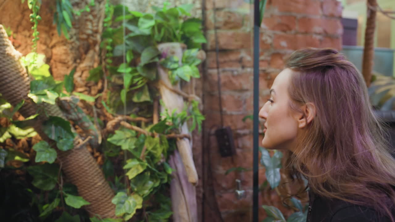 Blond woman with wavy hair bends forward curiously while observing lush green jungle-themed decoration filled with vines and branches, reaching hand forward in fascination under warm ambient light