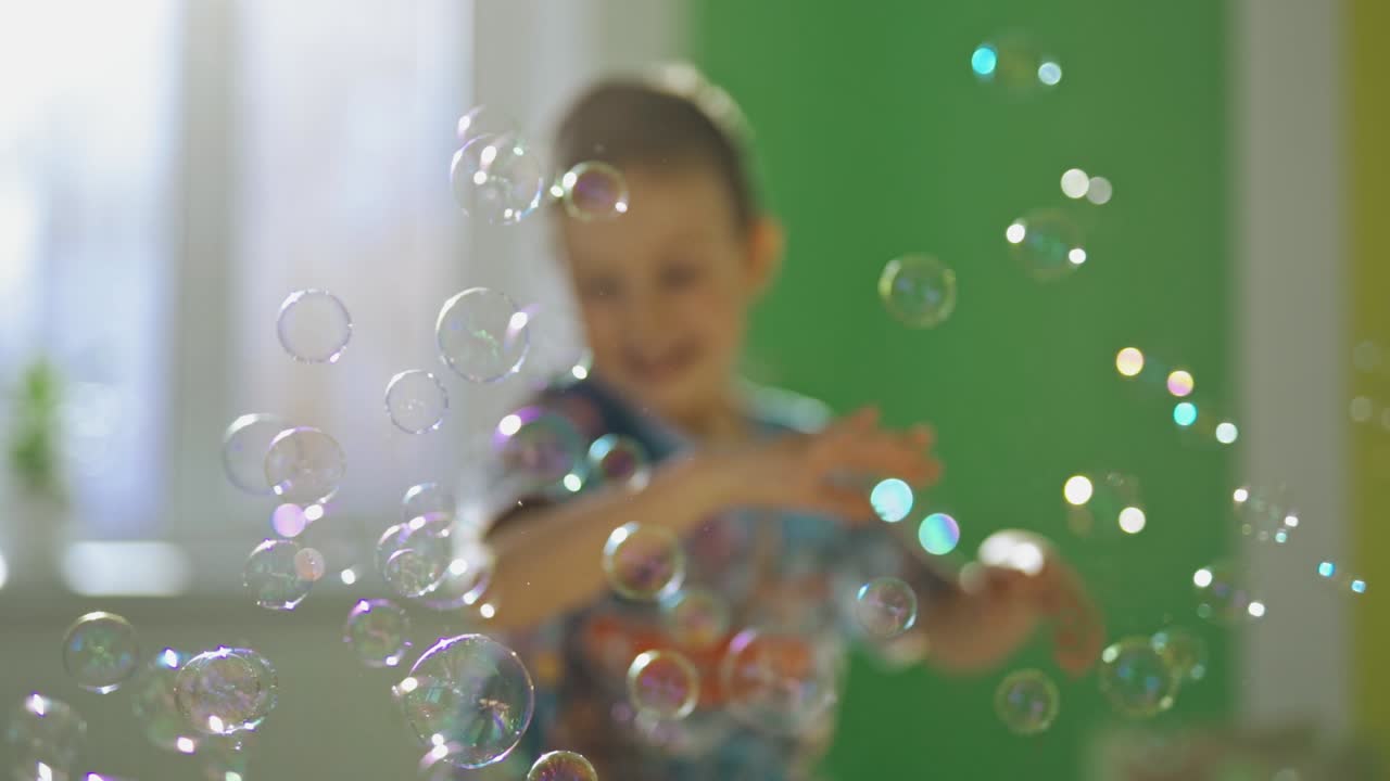 Round bubbles are flying inside the room and a boy catching and bursting them. Happy kid is playing with colorful soap bubbles at home.