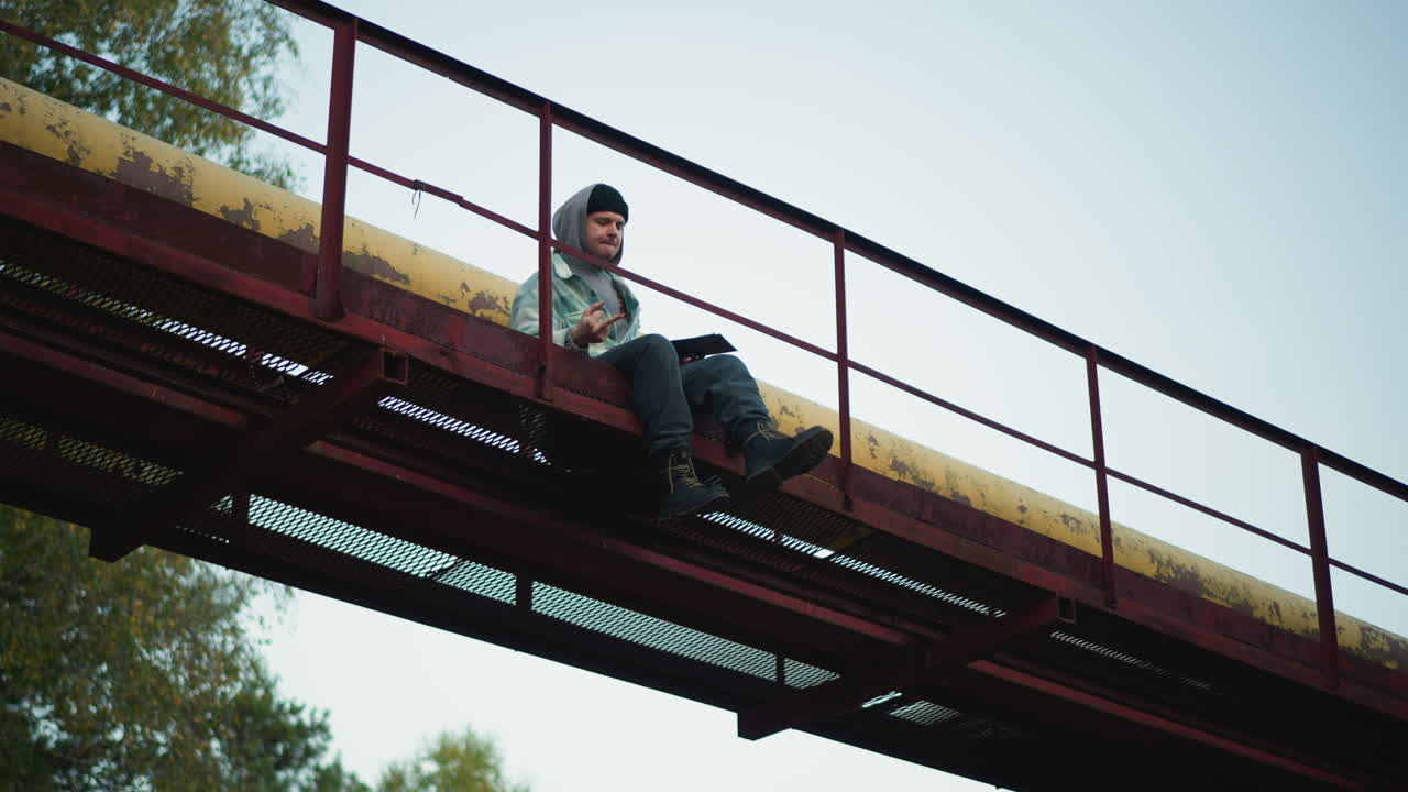 Caucasian hiker sitting on red catwalk overlooking treetops, backpack beside him, overcast sky, pensive mood, industrial metal structure and railing, quiet urban adventure and introspective pause