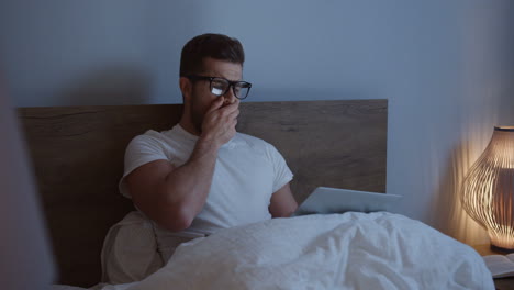 Tired-sleepy-young-man-working-late-at-night-on-the-laptop-computer-in-the-bed,-then-taking-off-his-glasses-and-yawning