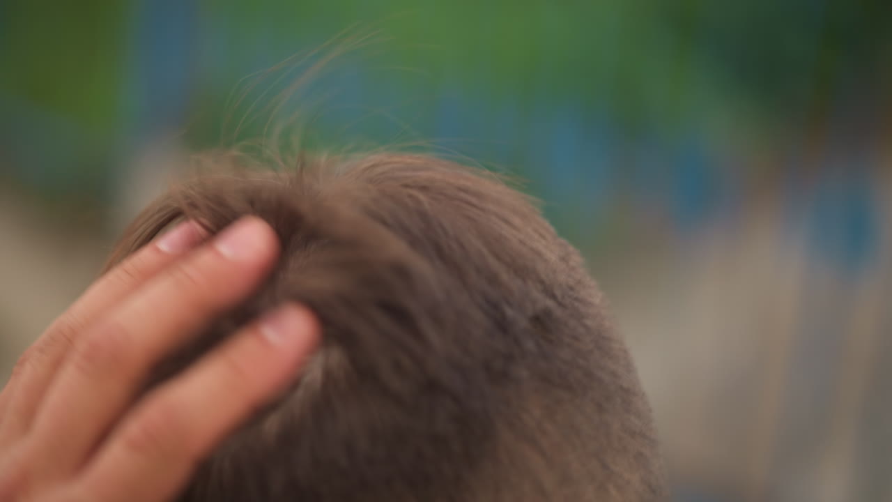 흐릿한 공원 풍경과 부드러운 햇살 아래서 초조한 듯 캐주얼한 복장을 한 남자가 머리를 매만지는 클로즈업 장면, 남성 개인이 야외에서 헤어스타일을 다듬고 있어요.