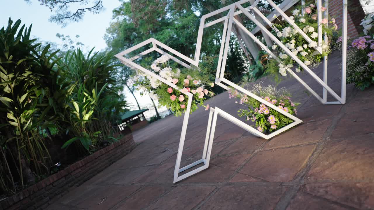 Angled right tracking shot of a white wire "XV" (15) structure decorated with flowers at the entrance of a Quinceañera party outdoors on a sunny afternoon