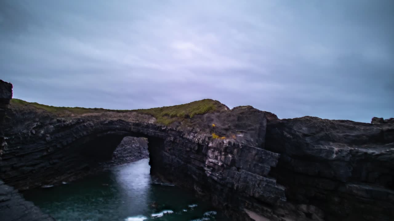 Timelapse of blue hour waves as ocean water crashes and rises below natural sea rch at Loop Head Kilkee, light fades over rugged coastal cliffs as clouds race in sky
