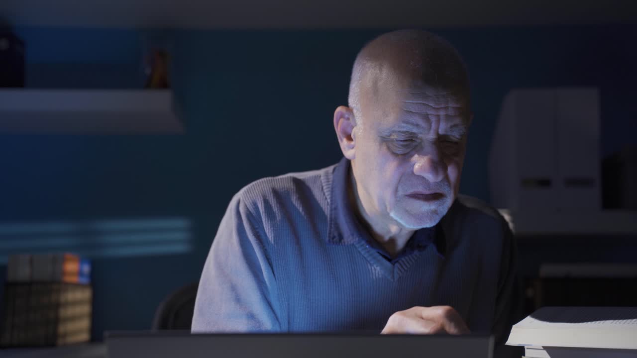 Old man reading a book, doing research, gaining knowledge using laptop in his working office.