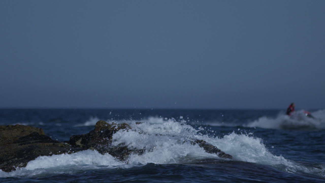 Ocean waves crash against a rock in the sea in Spain whilst a jet skier cruises past in the background, 4K slow motion