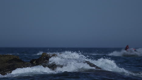 ocean waves crash against a rock in the sea in spain whilst a jet skier cruises past in the background, 4k slow motion