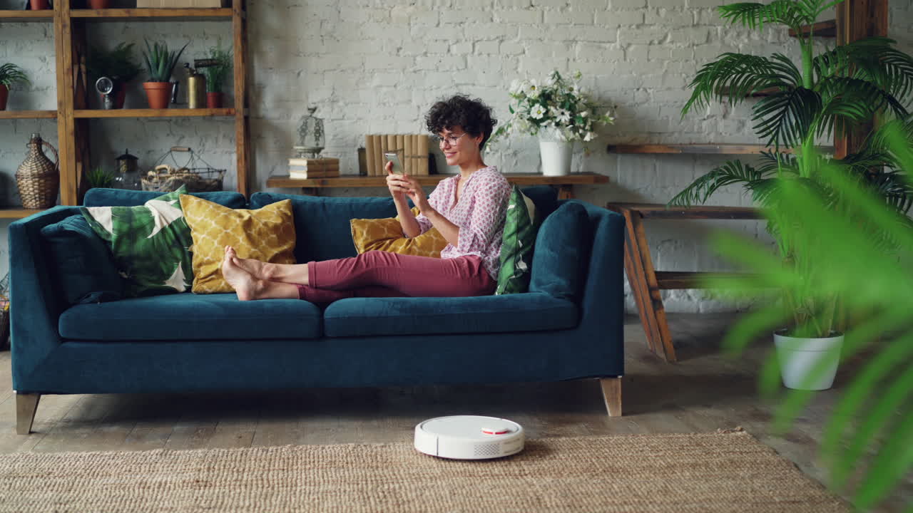 Woman relaxing on a sofa using smartphone while robotic vacuum cleaner cleans the floor