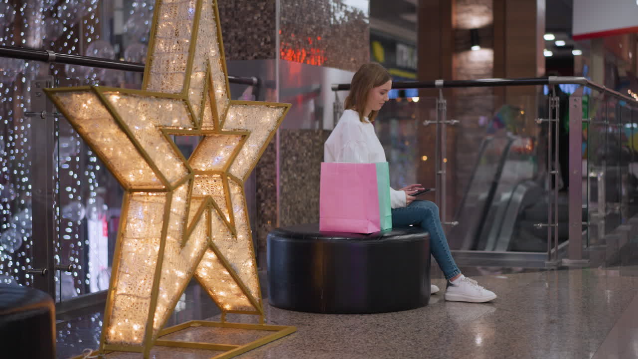 mujer joven sentada en un banco circular en un centro comercial usando su teléfono, rodeada de bolsas de compras, estrella iluminada decorativa y pantalla de luz brillante