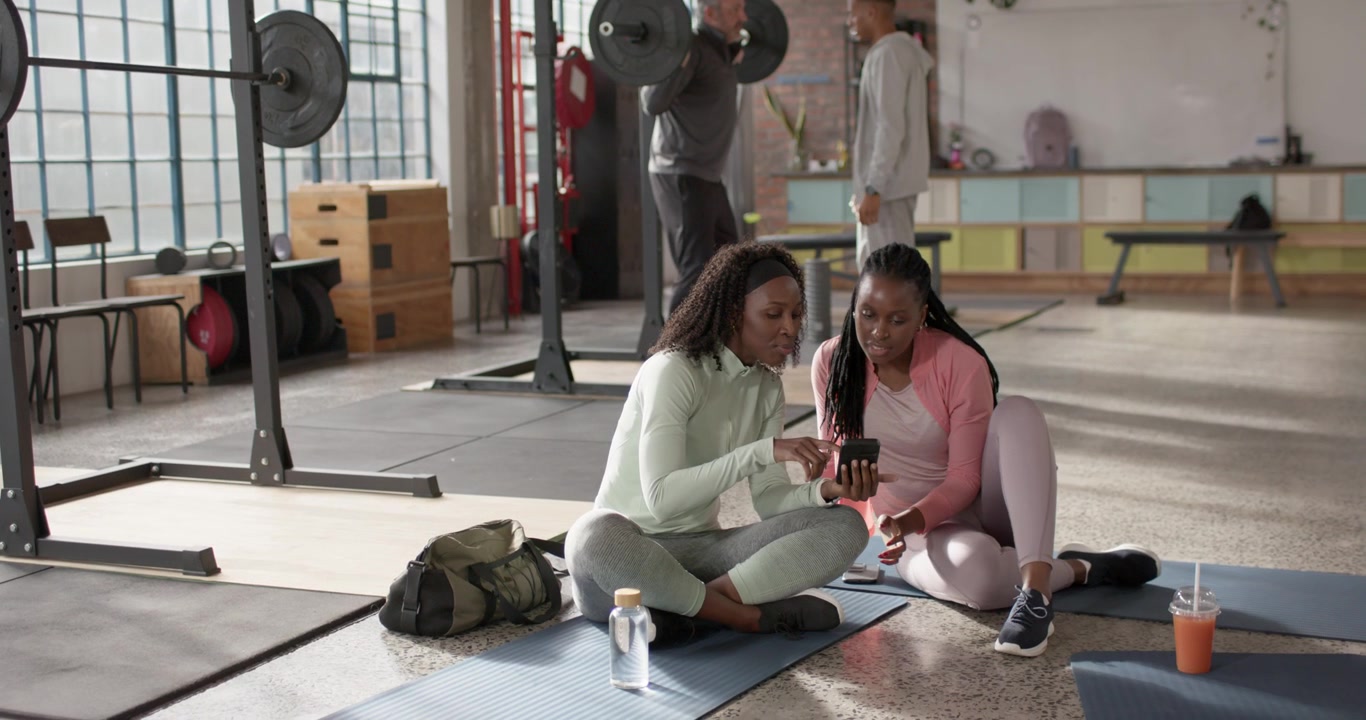 African American women talking, tapping smartphone, showing screen in gym while man adjusting bar