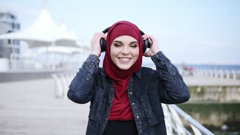 Slow-motion-footage-of-a-young-attractive-girl-with-hijab-on-her-head-putting-headphones-on,-smiling-and-enjoying-the-music-while-walking-near-sea-side