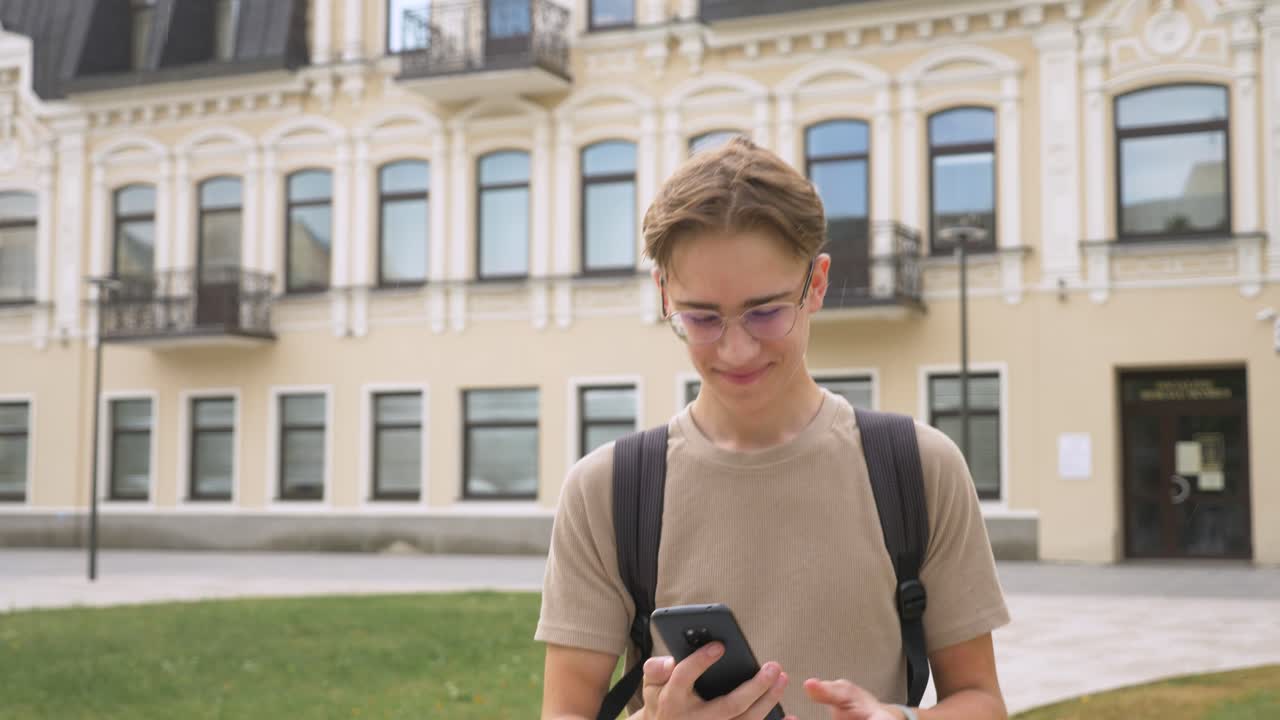 Primo piano di un adolescente felice che esce da scuola dopo le lezioni e sorride mentre messaggia con il suo telefono.