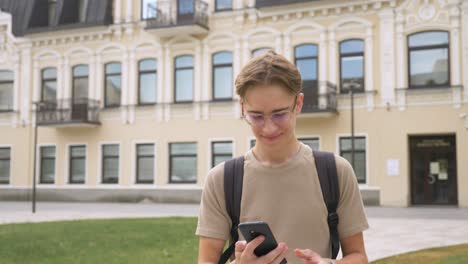 primer plano de un adolescente feliz caminando de la escuela después de las lecciones y sonriendo mientras envía mensajes de texto con su teléfono