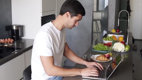 Der-Brünette-Junge-Mann-Sitzt-Morgens-Zu-Hause-An-Der-Küchentheke-Und-Arbeitet-Mit-Dem-Laptop.-Mann-Checkt-E-Mails,-Soziale-Medien-Im-Internet,-Tippt,-Während-Er-Einen-Tee-Aus-Einer-Tasse-Nippt