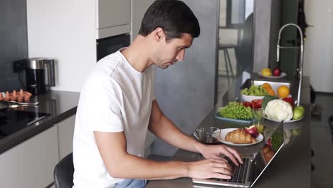 hombre moreno sentado y usando una computadora portátil trabajando por la mañana en casa en el mostrador de la cocina. hombre chequeando correos electrónicos, redes sociales, internet, escribiendo mientras toma un té de una taza