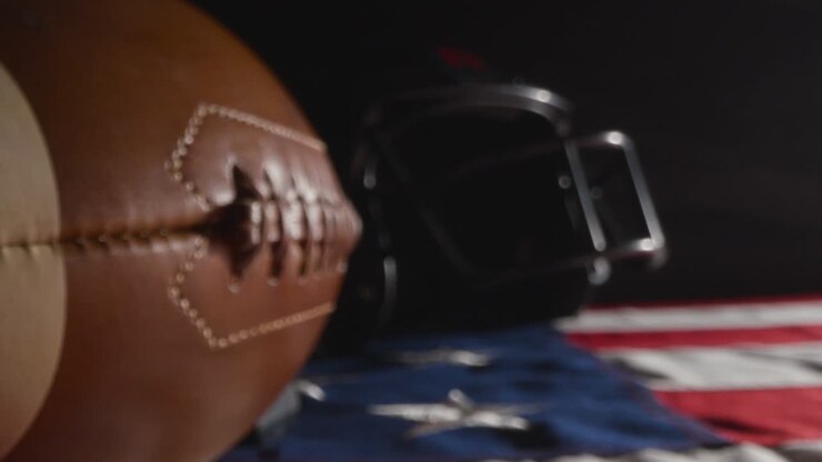 Toma de estudio con iluminación tenue de un balón de fútbol americano con casco sobre la bandera de barras y estrellas 2