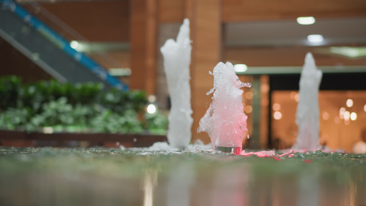 타일로 된 기둥, 에스컬레이터, 울창한 초록색으로 둘러싸인 현대 쇼핑몰 환경에서 다채로운 불빛으로 조명된 활기찬 하천으로 된 실내 물 분수