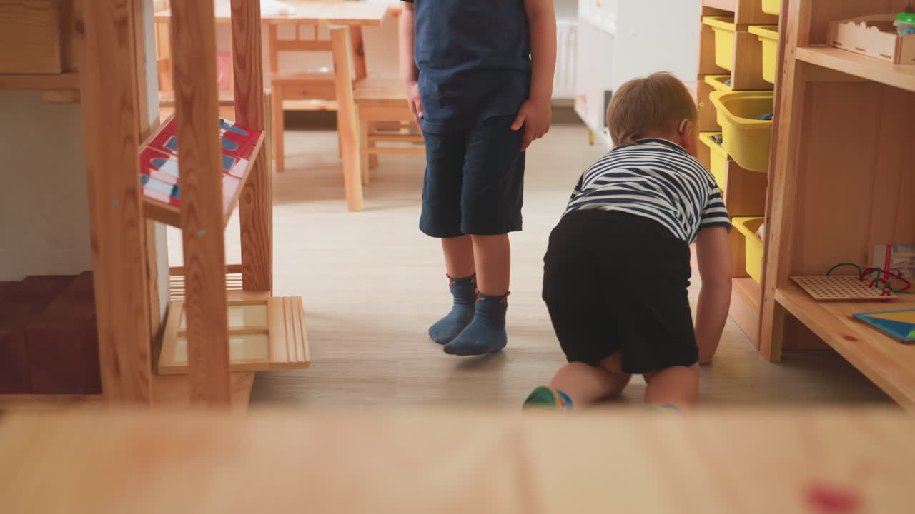 Besonderes Kind mit Hörgerät im gestreiften Hemd krabbelt auf dem Klassenzimmerboden und dreht den Kopf mit Strabismus, während ein Junge in Blau dahinter steht und die Szene in einer Lernumgebung mit gelben Aufbewahrungsboxen beobachtet