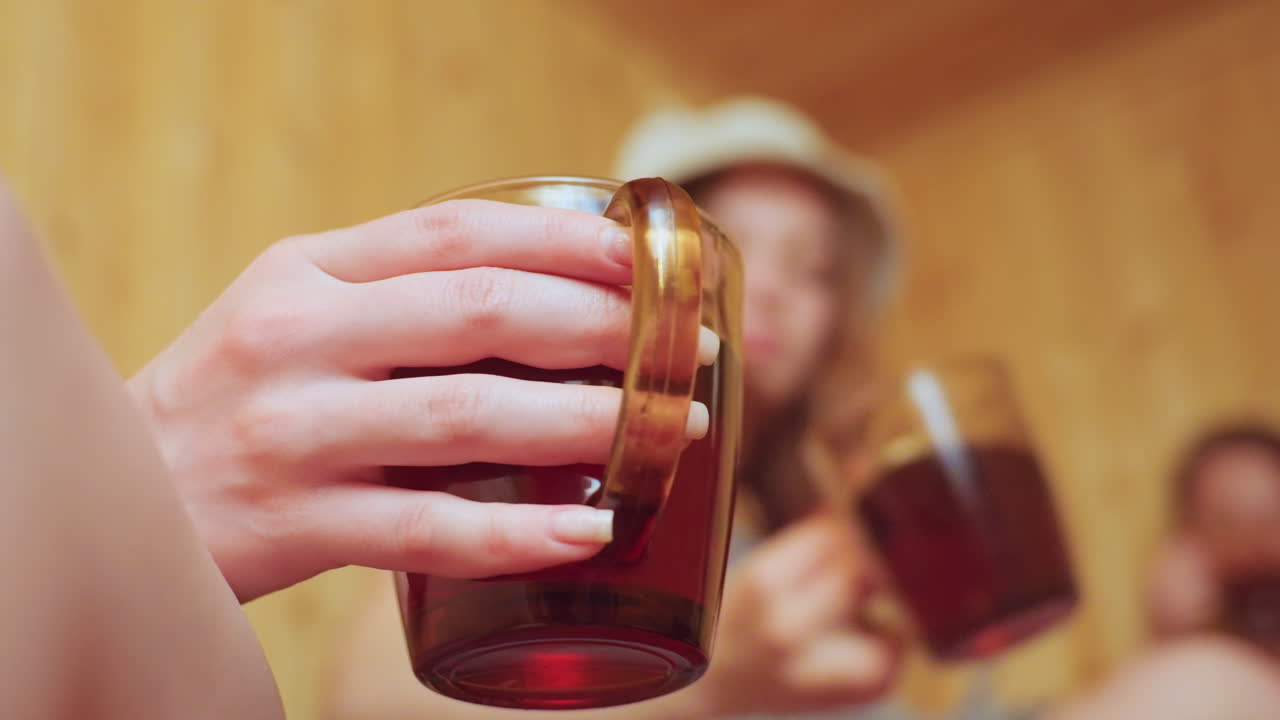 Close up of delicate hand holding transparent mug filled with native tea while in blurred background two women wrapped in towels make joyful toast inside cozy wooden sauna