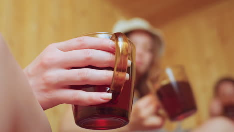close up of delicate hand holding transparent mug filled with native tea while in blurred background two women wrapped in towels make joyful toast inside cozy wooden sauna