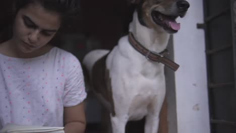 Una-Joven-Dibujando-Con-Un-Lápiz-En-Un-Cuaderno-De-Bocetos-Con-Un-Perro-Parado-A-Su-Lado-Sentado-En-La-Puerta