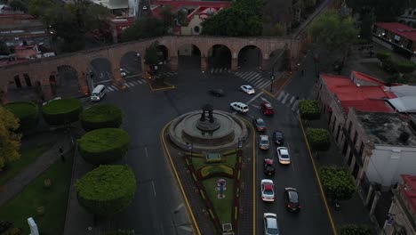 Drone-view-of-the-iconic-Tarascas-Fountain-in-Morelia,-Michoacán,-with-cars-circling-the-roundabout-and-the-city's-famous-aqueduct-arches