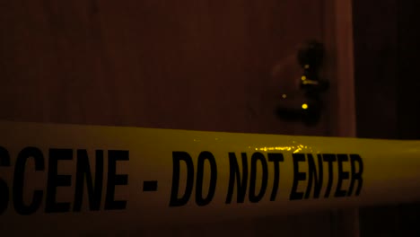 yellow colored crime scene do not enter police caution tape ribbon pulled in front of a wooden brown apartment or a house door with its chrome handle visible in the background. dark scene during night