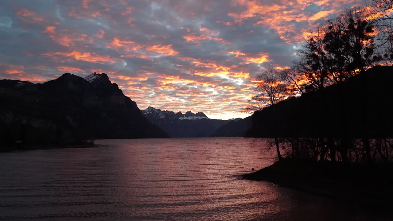 Ondulações na água refletem as cores vibrantes do nascer do sol nas nuvens, com silhuetas de árvores e montanhas em Walensee, Suíça.