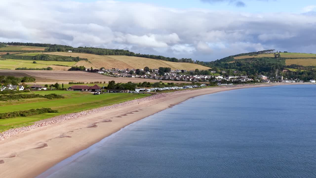 Drone camera smoothly pans above tranquil Rosemarkie shoreline, revealing village, hills, and summer landscape