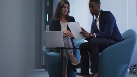 Diverse-businessman-and-businesswoman-discussing-documents-and-using-laptop-in-modern-office