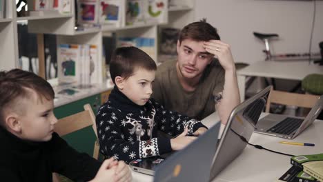 Young-male-attactive-teacher-in-his-20's-explaining-something-to-two-little-boys,-teaching-them-how-to-use-laptop.-Programming-class.-Educational-process.