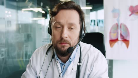 close-up view of senior male doctor with headphones sitting at desk speaking at camera and giving medical advice about coronavirus in hospital office