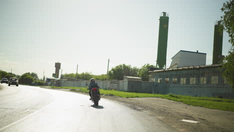 back view of a female biker, her clothes fluttering in the wind as she rides down the road, cars are seen coming in the opposite direction, with a factory building and trees visible by the right