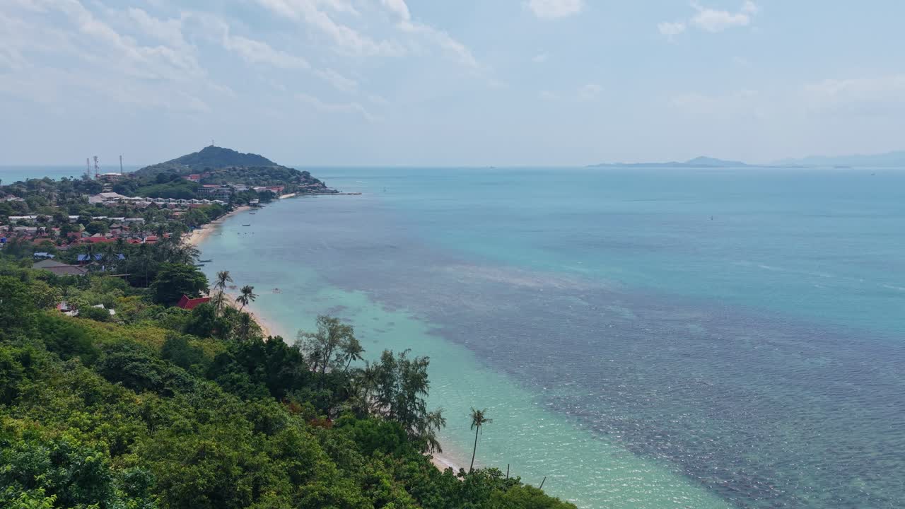 Cinematic aerial movement over lush tropical hillside with turquoise sea and distant beach town