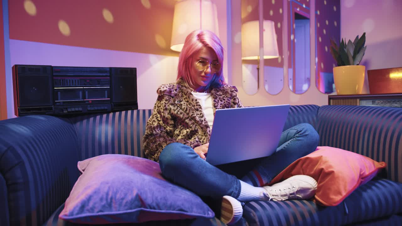 Happy young Caucasian stylish woman in sunglasses sitting on sofa and using laptop in vintage living room at home