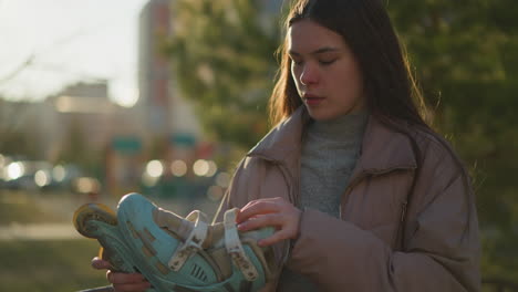 A young woman wearing a peach jacket and a gray sweater is seen adjusting the straps of her light blue rollerblades while standing in a park. The blurred background includes green trees