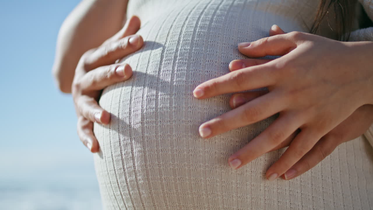 의 손이 배를 만지고 임신한 여성이 빛의 자연에 가까이 서 있습니다.