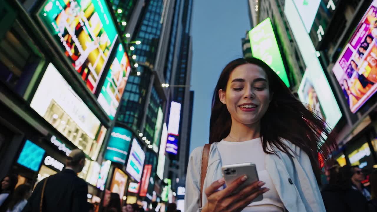 Low-angle shot of a smiling woman using her phone in a vibrant cityscape with bright billboards