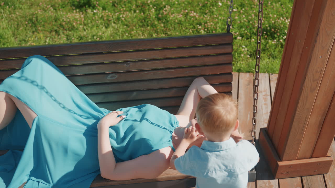 Family enjoying pleasant park afternoon. Woman and child share joyful moments on bright weekend day. Relaxed mother watches toddler explore wooden swing on sunny family park afternoon