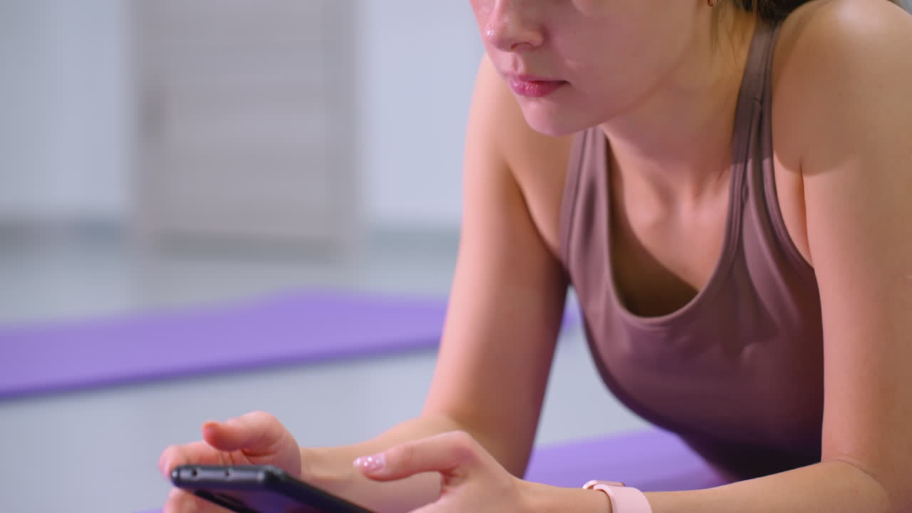 mujer enfocada acostada operando un teléfono inteligente con una alfombra de yoga púrpura cerca, un primer plano captura su expresión concentrada, postura relajada y iluminación suave