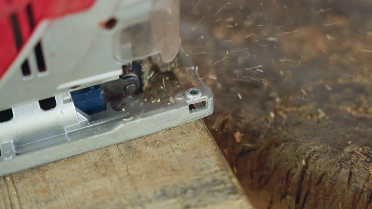 パレットの端をトリミングするダイナミックなジグソー。赤い工具ハウジングから素早く切りくずが排出され、細かい削りくずが舞う。粗い木目に接触する刃のクローズアップ、木のダボとトリミングされた端が現れる。活気ある作業場。